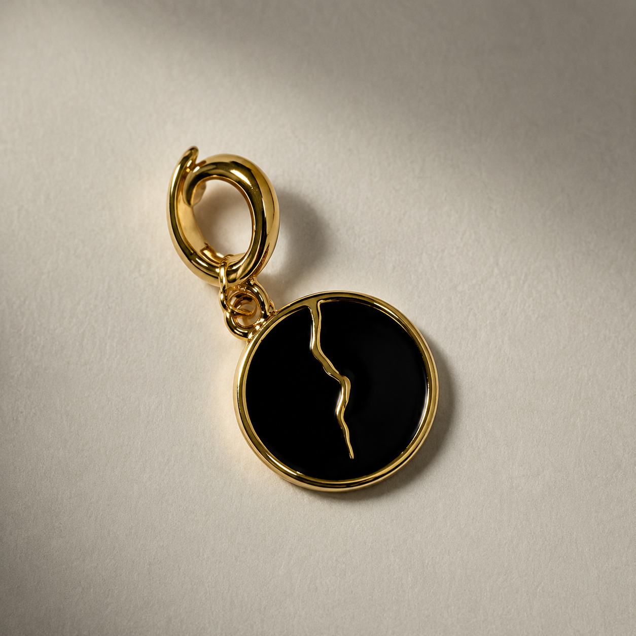 a Make Savage circular gold toned black enamel charm with a gold-toned crack representing the seeking of mastery and its inherent impossibility.