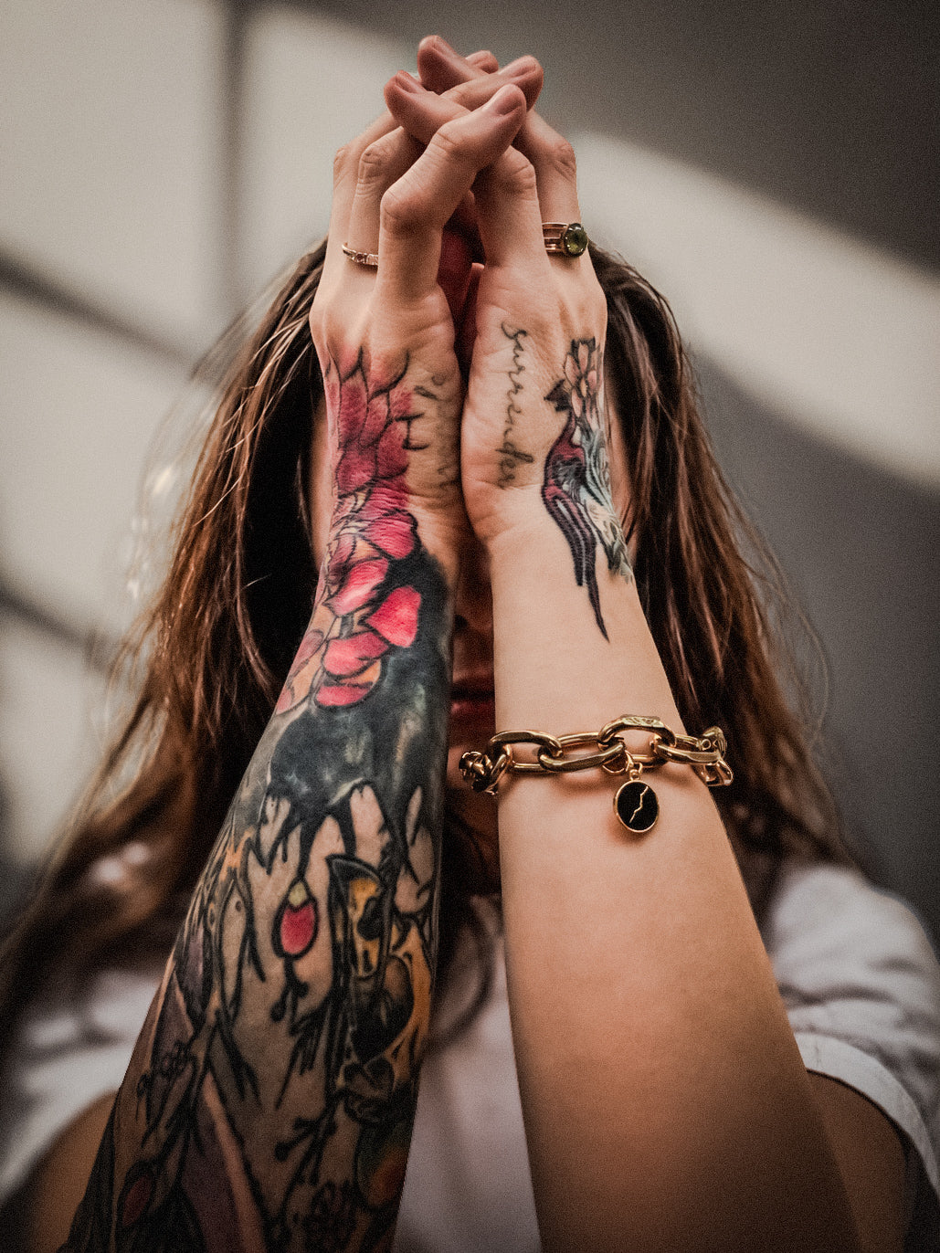 Close-up of two tattooed forearms and hands clasped together.. A gold chain bracelet with a Make Savage Black Enamel Charm is worn on a bracelet on one wrist.